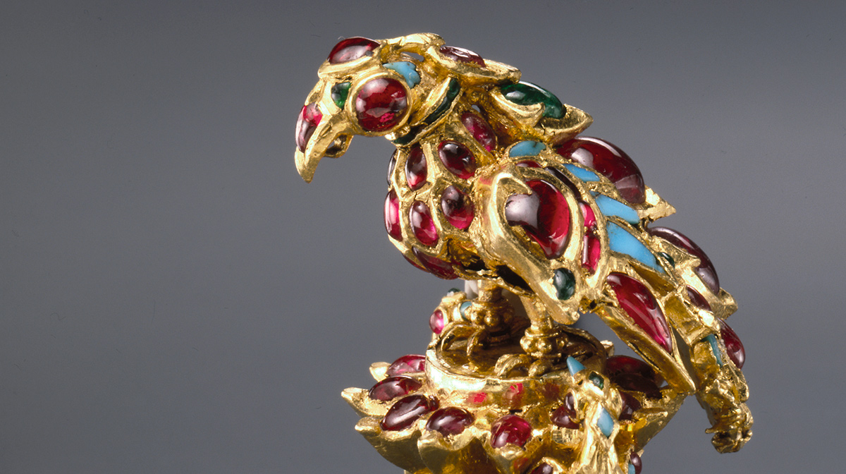 A ring with a bird of prey on top from the al-Sabah Collection. The ring is 17th century gold and covered with blue and red encrusted jewels including rubies, emeralds, and turquoise.